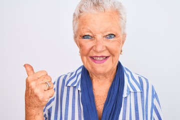 Senior grey-haired woman wearing blue striped shirt standing over isolated white background pointing and showing with thumb up to the side with happy face smiling