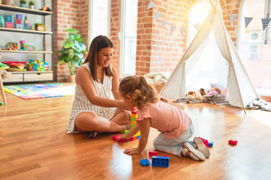 Beautiful teacher and blond toddler girl building tower using plastic blocks at kindergarten