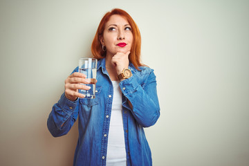 Young beautiful redhead woman drinking glass of water over white isolated background serious face...