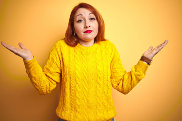 Beautiful redhead woman wearing winter sweater standing over isolated yellow background clueless...