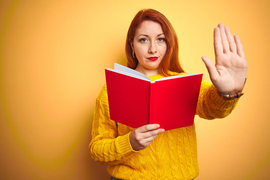 Young redhead teacher woman reading red book over yellow isolated background with open hand doing stop sign with serious and confident expression, defense gesture