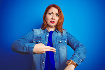Young beautiful redhead woman wearing denim shirt standing over blue isolated background In hurry...
