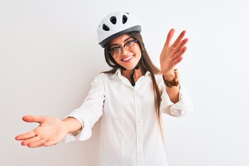 Beautiful businesswoman wearing glasses and bike helmet over isolated white background looking at the camera smiling with open arms for hug. Cheerful expression embracing happiness.