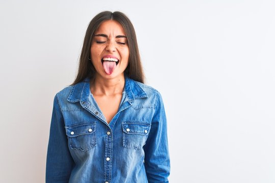 Young beautiful woman wearing casual denim shirt standing over isolated white background sticking tongue out happy with funny expression. Emotion concept.