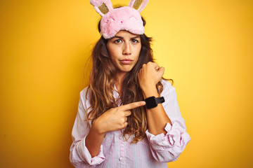 Young woman wearing pajama and sleep mask standing over yellow isolated background In hurry...