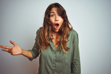 Young beautiful woman wearing green shirt standing over grey isolated background In shock face,...