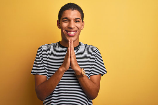 Young handsome arab man wearing navy striped t-shirt over isolated yellow background praying with hands together asking for forgiveness smiling confident.