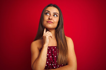 Young beautiful woman wearing casual t-shirt standing over isolated red background with hand on chin thinking about question, pensive expression. Smiling with thoughtful face. Doubt concept.