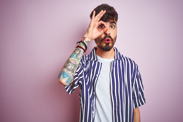 Fototapeta premium Young man with tattoo wearing striped shirt standing over isolated pink background doing ok gesture shocked with surprised face, eye looking through fingers. Unbelieving expression.