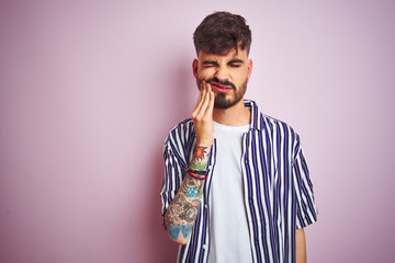 Fototapeta premium Young man with tattoo wearing striped shirt standing over isolated pink background touching mouth with hand with painful expression because of toothache or dental illness on teeth. Dentist concept.