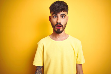 Young man with tattoo wearing t-shirt standing over isolated yellow background afraid and shocked with surprise expression, fear and excited face.