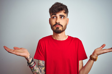 Young man with tattoo wearing red t-shirt standing over isolated white background clueless and...