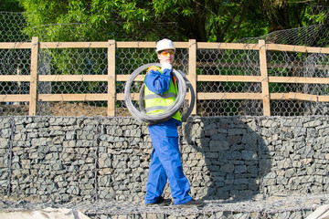 Obraz na płótnie Canvas a worker in a white helmet, against a background of a wooden fence and building stones, carries a wire on his shoulder