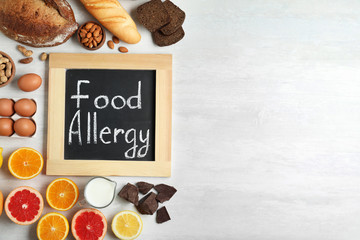 Chalkboard with words FOOD ALLERGY and different products on white table, flat lay. Space for text