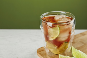 Glass of tasty ice tea with lime on white wooden table against green background, space for text