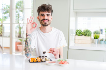 Fototapeta na wymiar Young man eating asian sushi from home delivery doing ok sign with fingers, excellent symbol