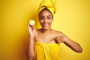 Afro woman wearing towel after shower holding slice lemon over isolated yellow background with...