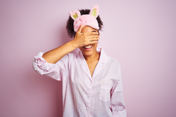 Young african american woman wearing pajama and mask over isolated pink background smiling and laughing with hand on face covering eyes for surprise. Blind concept.