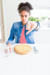 Young african american woman eating homemade cheese pizza with angry face, negative sign showing dislike with thumbs down, rejection concept