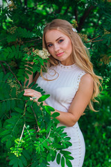 Portrait of a charming blond woman wearing beautiful white dress standing next to rowan tree.