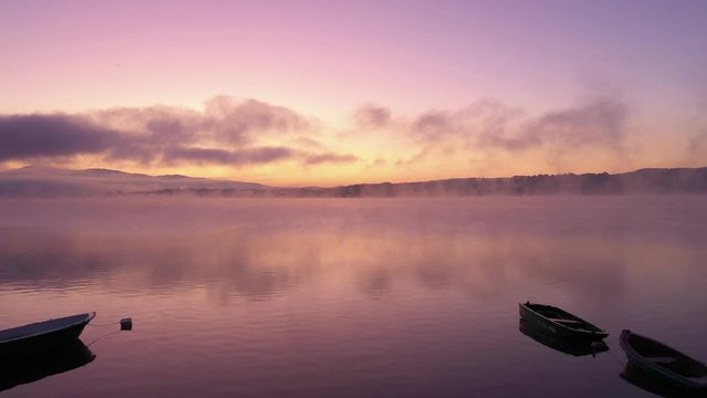 Dawn at a lake with amazing colours, anglers boats by a shore, flying through a mist floating over a water surface. Aerial dolly shot