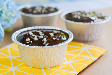 Dark chocolate cake with chocolate chips topping in round foil cup on yellow tablecloth and wooden table. Copy space for your text. Selective focus.