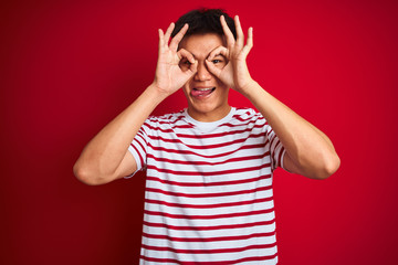 Young asian chinese man wearing striped t-shirt standing over isolated red background doing ok gesture like binoculars sticking tongue out, eyes looking through fingers. Crazy expression.