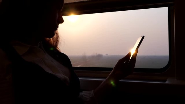 Passenger woman sitting with smartphone against window, ride at high speed train. Evening sun light shine from outside and reflex flick on the screen of mobile device, silhouetted shot