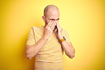 Young bald man with beard wearing casual striped t-shirt over yellow isolated background rubbing...
