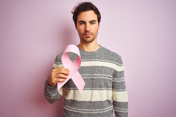 Young handsome man holding cancer ribbon standing over isolated pink background with a confident...