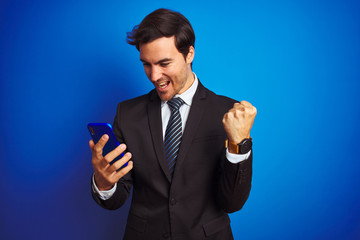 Young handsome businessman using smartphone standing over isolated blue background screaming proud...
