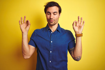 Young handsome man with tattoo wearing casual shirt standing over isolated yellow background relax and smiling with eyes closed doing meditation gesture with fingers. Yoga concept.