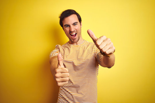 Young handsome man with tattoo wearing striped t-shirt over isolated yellow background approving doing positive gesture with hand, thumbs up smiling and happy for success. Winner gesture.