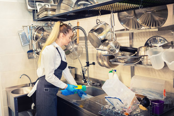 Young woman worker washing dishes using sponge in restaurant sink at end of working day