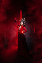 beautiful fashion witch woman with horns in the form of tree roots in a long luxurious dress on the background of a huge gate with red smoke. Halloween concept
