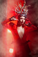 beautiful fashion witch woman with horns in the form of tree roots in a long luxurious dress on the background of a huge gate with red smoke. Halloween concept