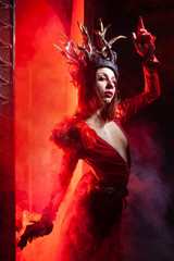 beautiful fashion witch woman with horns in the form of tree roots in a long luxurious dress on the background of a huge gate with red smoke. Halloween concept