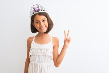 Beautiful child girl wearing princess crown standing over isolated white background smiling with happy face winking at the camera doing victory sign. Number two.