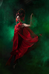 strict and scary girl in a fashionable long red dress and a voluminous hat on the background of green smoke in the Studio at night alone. Halloween concept.