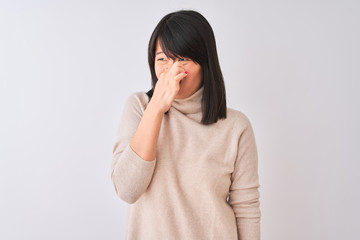 Young beautiful chinese woman wearing turtleneck sweater over isolated white background smelling something stinky and disgusting, intolerable smell, holding breath with fingers on nose