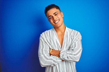 Young brazilian man wearing striped shirt standing over isolated blue background happy face smiling with crossed arms looking at the camera. Positive person.