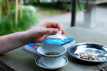 Close-up of cup of tea on the table in China, a hand raising the lid of the cup,  background whit touristic guide and seeds out of focus
