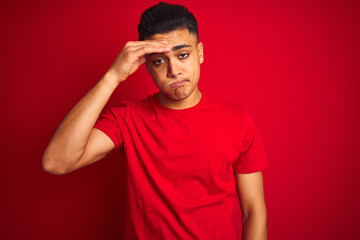 Young brazilian man wearing t-shirt standing over isolated red background worried and stressed about a problem with hand on forehead, nervous and anxious for crisis