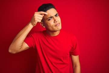 Fototapeta premium Young brazilian man wearing t-shirt standing over isolated red background pointing unhappy to pimple on forehead, ugly infection of blackhead. Acne and skin problem