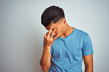 Young brazilian man wearing blue t-shirt standing over isolated white background tired rubbing nose and eyes feeling fatigue and headache. Stress and frustration concept.