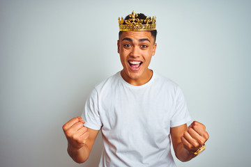 Young brazilian man wearing king crown standing over isolated white background celebrating surprised and amazed for success with arms raised and open eyes. Winner concept.