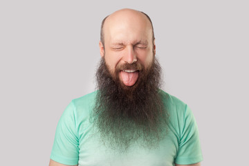 Portrait of funny middle aged bald man with long beard in light green t-shirt standing with closed eyes and tongue out. indoor studio shot, isolated on grey background.