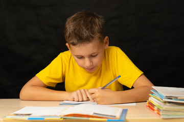 The boy solves homework, sitting at his desk. The boy writes homework with his left hand