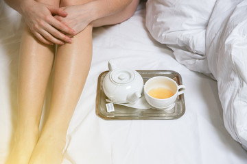 Woman is sitting in the bed. Beautiful slim legs. Girl with tea and cup on the tray. Morning at the hotel.