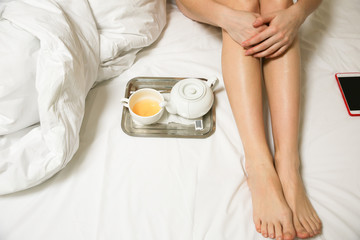 Woman is sitting in the bed. Beautiful slim legs. Girl with tea and cup on the tray. Morning at the hotel.